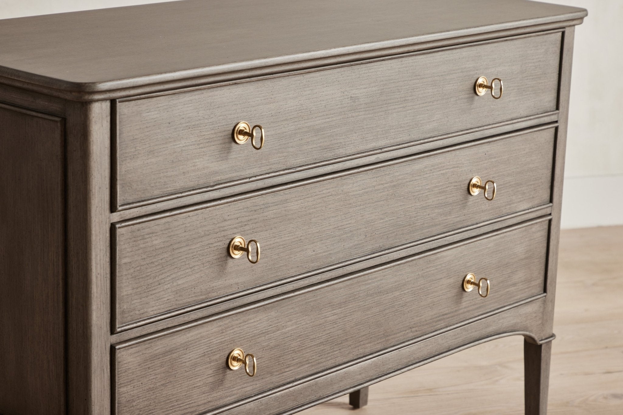 Brown wooden dresser with three drawers and round knobs, standing on tapered legs, in a minimal room.