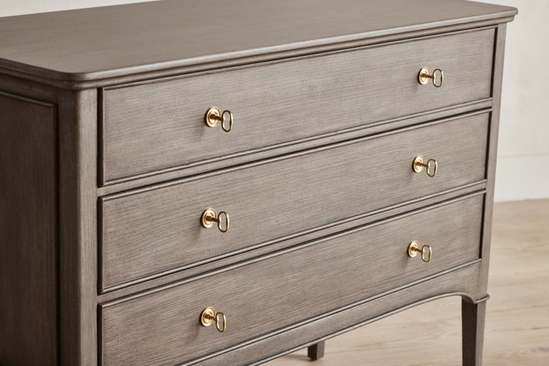 Dark gray-finished dresser with four drawers and gold knobs, standing on tapered legs.
